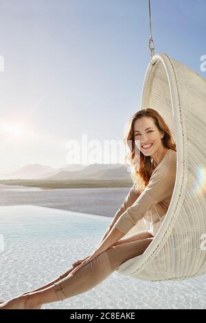 Smiling woman relaxing in hanging chair above swimming pool Stock Photo