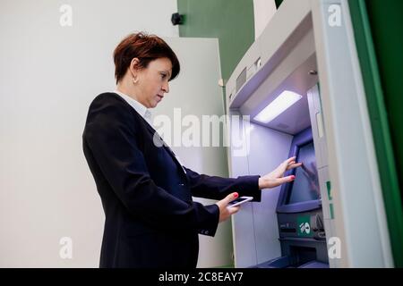 Businesswoman holding smart phone using ATM while standing by wall Stock Photo