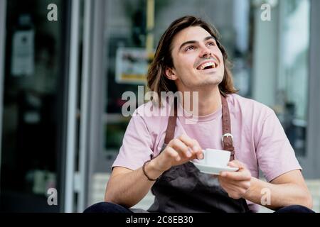 Male owner holding coffee cup laughing while sitting outside cafe Stock Photo