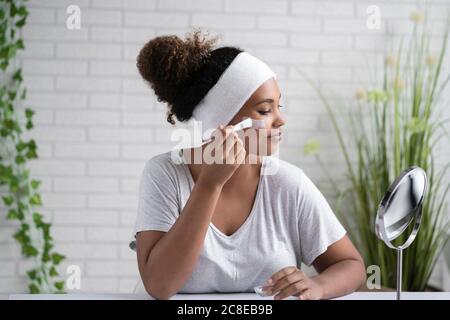 Young woman wearing headband applying facial mask while looking in mirror at home