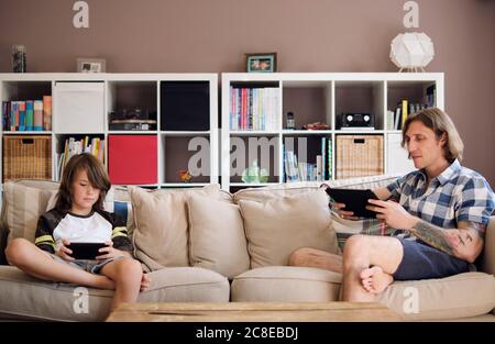 Father and son using technologies while sitting on sofa at home Stock Photo