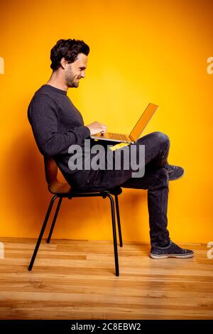 Smiling man using laptop while sitting on chair by yellow background Stock Photo