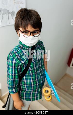 Boy wearing mask holding skateboard while standing on steps in school Stock Photo