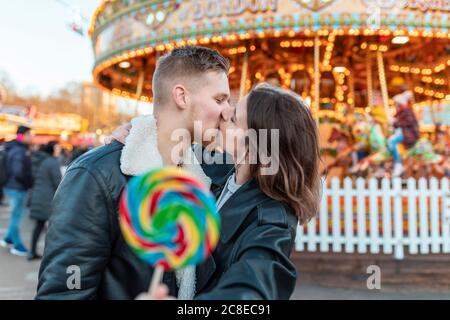 Young couple holding lollipop kissing while standing at amusement park