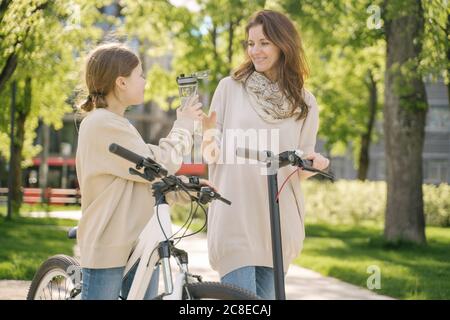 Girl sharing water with mother while standing in city park Stock Photo