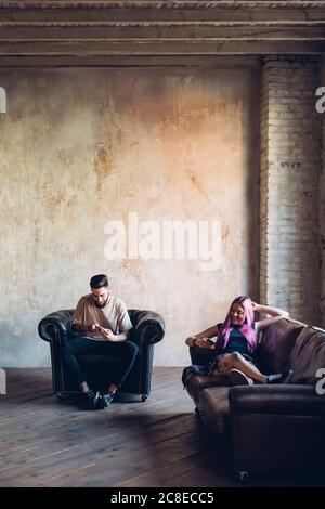 Young man and woman sitting on sofa and armchair in a loft using smartphones Stock Photo