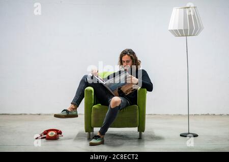 Young man reading book while relaxing on armchair against wall at home Stock Photo