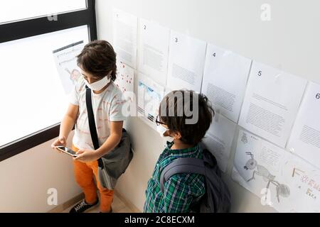 Boy wearing mask looking at friend using smart phone while standing in school Stock Photo