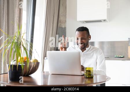 Portrait of happy man sitting at kitchen table during video chat showing victory sign Stock Photo