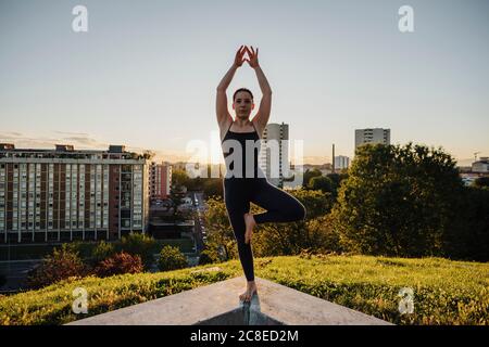 Young woman practicing yoga in tree pose on retaining wall in city during sunset Stock Photo