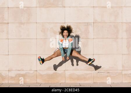 Excited young woman screaming while jumping against wall during sunny day Stock Photo
