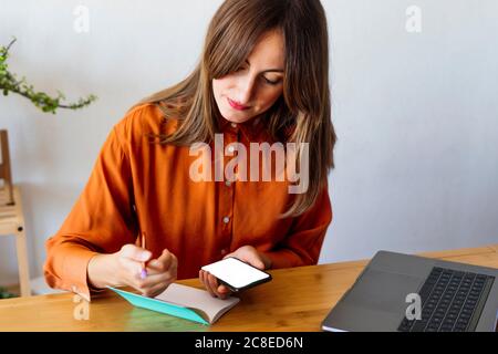 Female freelancer working at home sitting at desk with notepad and smartphone Stock Photo