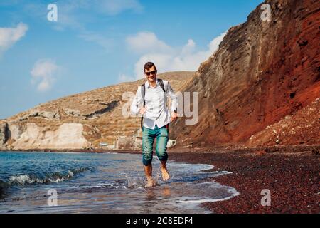 Happy tourist running on Red beach by sea on Santorini island barefooted. Man with backpack traveling Stock Photo