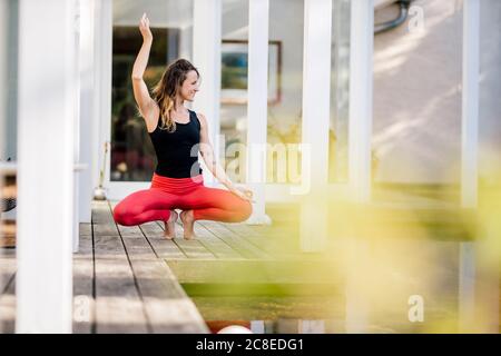 Woman with arm raised exercising while crouching on hardwood floor against house Stock Photo