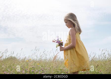 Blond little girl wearing yellow dress standing on a meadow with bouquet of picked field flowers Stock Photo
