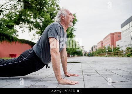 Senior man with mouth open doing push-ups on footpath in city Stock Photo