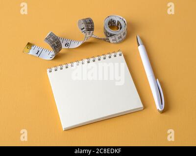 Open paper notepad with copy space, white pen and roll of measuring tape with centimeters and inches on a yellow textured background. Copy space. Stock Photo