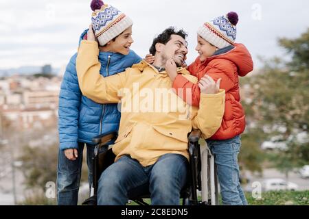 Loving father embracing sons while sitting on wheelchair in park Stock Photo