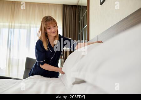 Smiling chambermaid arranging pillows on bed in hotel room Stock Photo