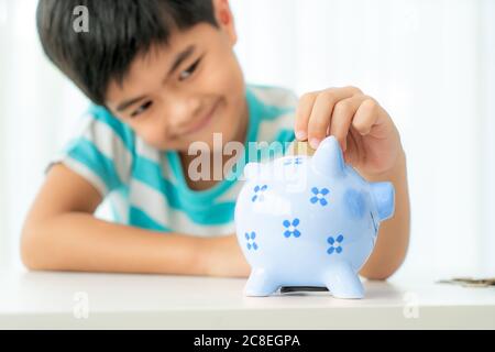 Little Asian boy insert a coin into blue piggy bank in white table at living room at home for the kid saving, investment money for his business future