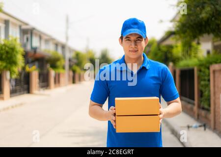 Asian delivery young man in blue uniform smile and holding pile of cardboard boxes in front house village with copy space. Advertising, Business, Tran Stock Photo