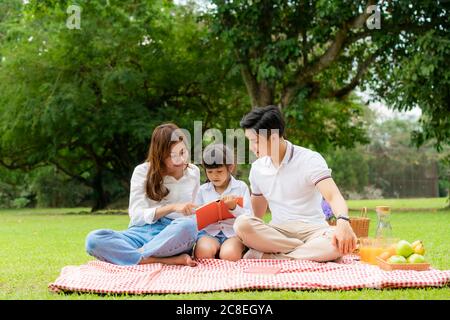 Asian teen family happy holiday picnic moment in the park with father, mother and daughter reading book togerter in green garden with fruit and food. Stock Photo