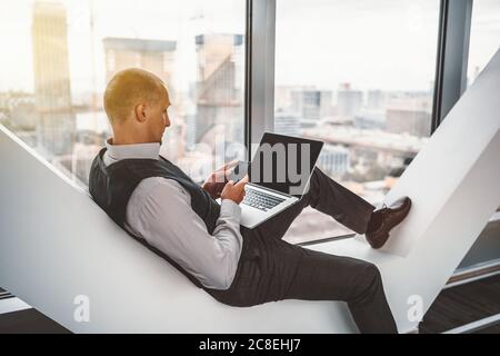 Hispanic mature man entrepreneur is relaxing on a modern white bench and using his smartphone, with the laptop on his knees, indoors of an office Stock Photo