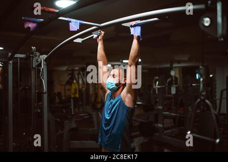 Muscular young fitness man with medical mask doing exercises on horizontal bar in a gym club. COVID 19 coronavirus protection Stock Photo