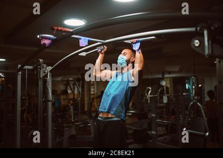 Muscular young fitness man with medical mask doing exercises on horizontal bar in a gym club. COVID 19 coronavirus protection Stock Photo