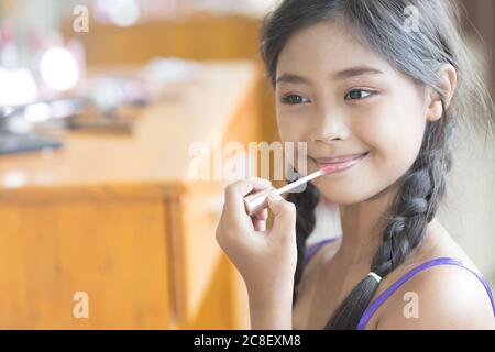 A cute long-haired Asian schoolgirl is using lipstick to apply lip makeup during a makeup lesson. Stock Photo