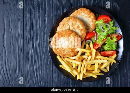 turkey burgers with lettuce tomato salad and chunky chips on a black plate on a wooden table, view from above, flatlay, free space for text Stock Photo