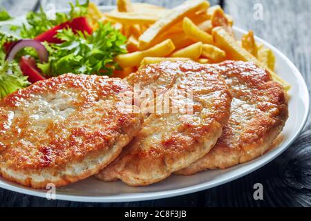 close-up of freshly fried three turkey burgers served with lettuce tomato salad and french fries on a white plate on a rustic wooden table, horizontal Stock Photo