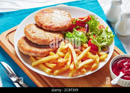 lettuce tomato salad and french fries served with freshly fried three turkey burgers on a white plate on a cutting board with tomato sauce in bowl, ho Stock Photo