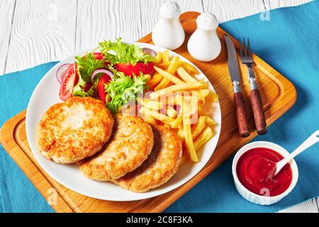 lettuce tomato salad and french fries served with freshly fried three turkey burgers on a white plate on a cutting board with tomato sauce in bowl, ho Stock Photo