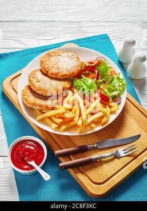 lettuce tomato salad and french fries served with freshly fried three turkey burgers on a white plate on a cutting board with tomato sauce in bowl, ve Stock Photo