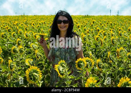 Cheerful brunette woman in sunflower field holding flower Stock Photo