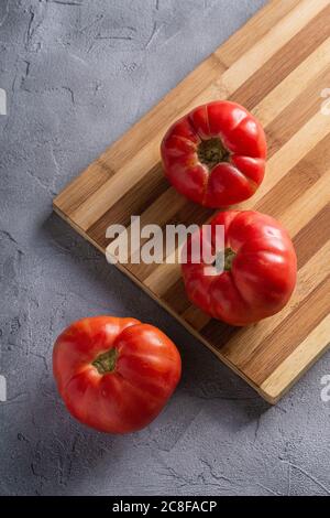 Three pink heirloom tomato vegetables, fresh red ripe tomatoes on wooden cutting board, vegan food, stone concrete background, angle view Stock Photo