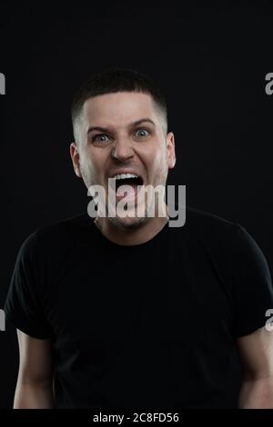 Emotional young man with round eyes, open mouth, showing astonishment and shock, isolated on black Stock Photo