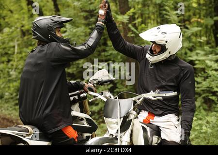 Young men in helmets sitting on motorbikes and giving high five while supporting each other during race in forest Stock Photo