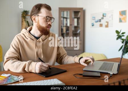 Serious creative young man in hoodie working on design project using drawing pad in home office Stock Photo