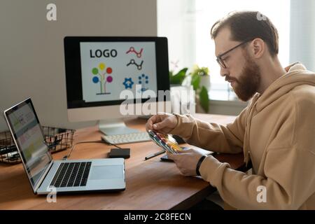 Serious bearded designer in eyeglasses sitting at desk with computers and viewing color swatch while working on logo design Stock Photo