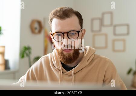Busy young bearded programmer in glasses working with laptop while analyzing pictures in home office Stock Photo