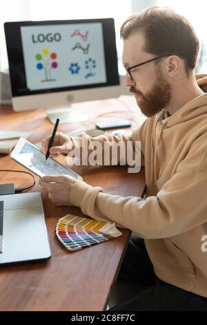 Concentrated bearded man in hoodie sitting at wooden table and using tablet while working on brand design Stock Photo