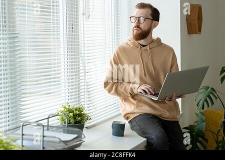 Pensive young bearded man in hoodie sitting on window-sill and holding laptop while thinking of design Stock Photo