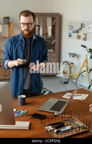 Focused creative young graphic designer with beard standing at table and looking at computer monitor while working with digitizer Stock Photo