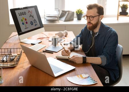 Concentrated young bearded man listening music in earphones and making sketch in notepad while working on digital drawing Stock Photo