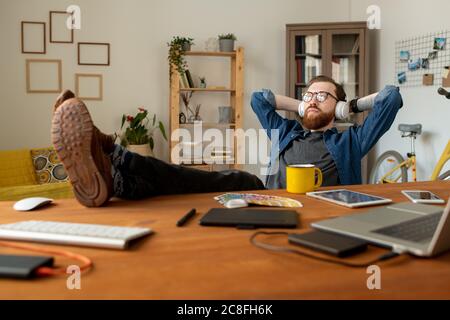 Dreamy young bearded man sitting in relaxed position and listening to music in headphones while thinking of new design project Stock Photo