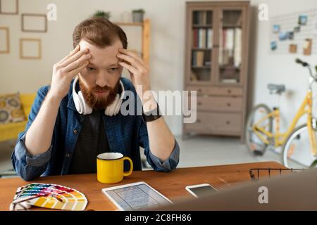 Puzzled young UI designer with beard sitting at table and holding head in hands while thinking of interface layout Stock Photo