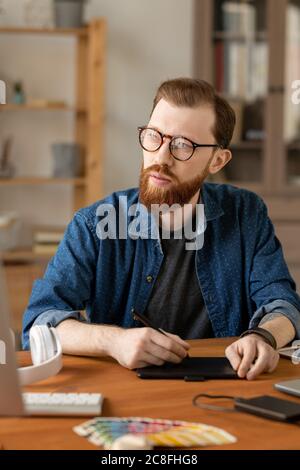 Hipster bearded man in eyeglasses sitting at table and using digitizer tablet while thinking of visual solution Stock Photo