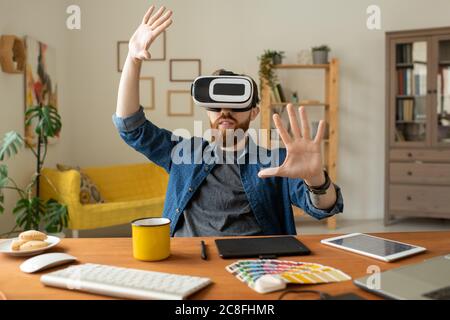 Content curious bearded man sitting at desk in home office and gesturing hands while testing new app in virtual reality goggles Stock Photo